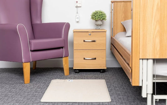 Wireless floor sensor mat on carpet between purple armchair and wooden hospital bed with bedside cabinet