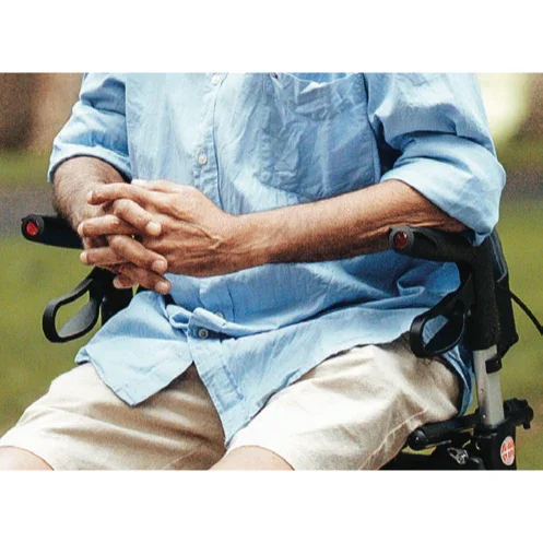 Elderly man wearing light blue shirt and beige shorts sitting in a wheelchair outdoors with hands clasped