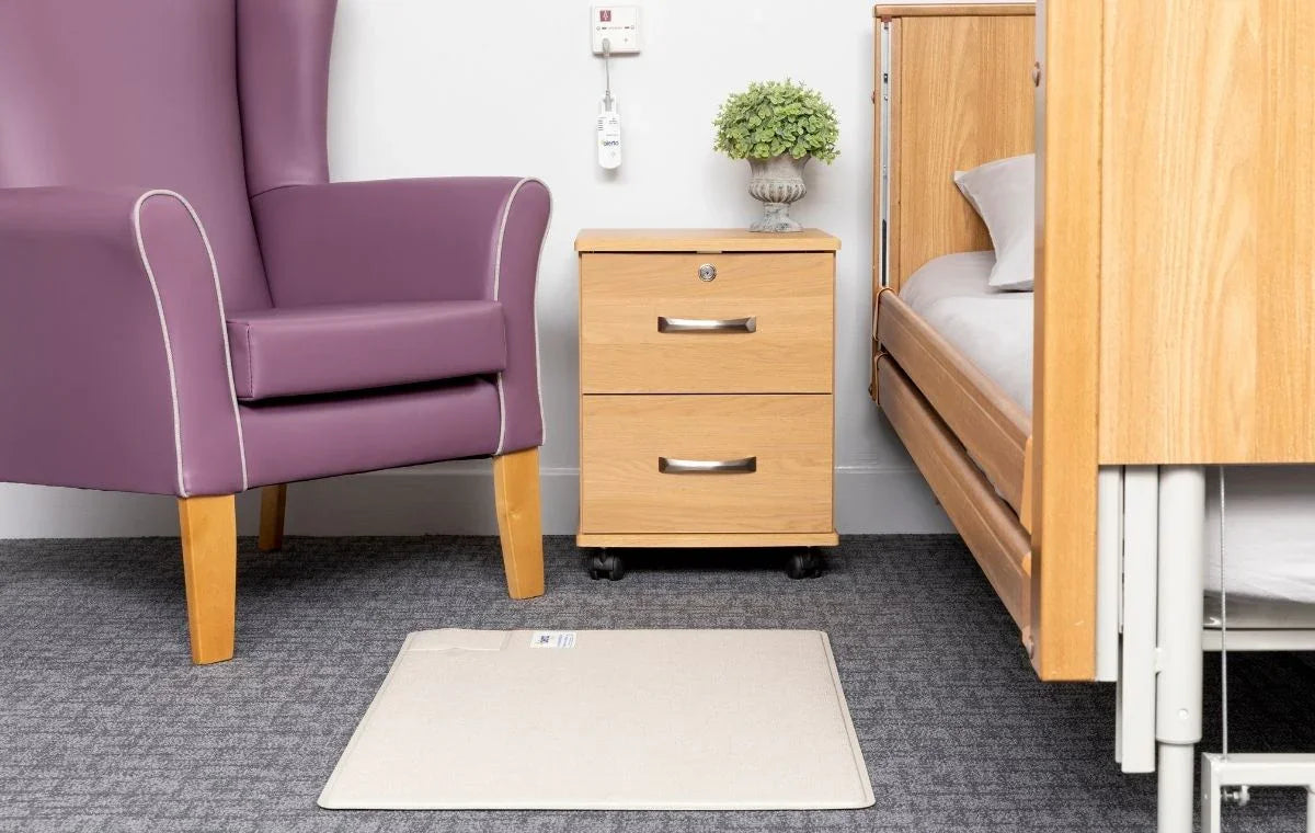 Wireless floor sensor mat on carpet between purple armchair and wooden hospital bed with bedside cabinet